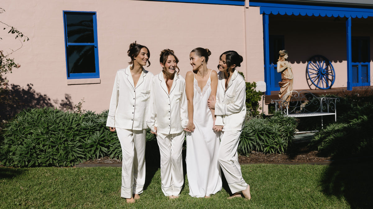 Bride in an ivory satin slip nightgown walking with three bridesmaids in matching ivory satin pyjama sets on the wedding morning