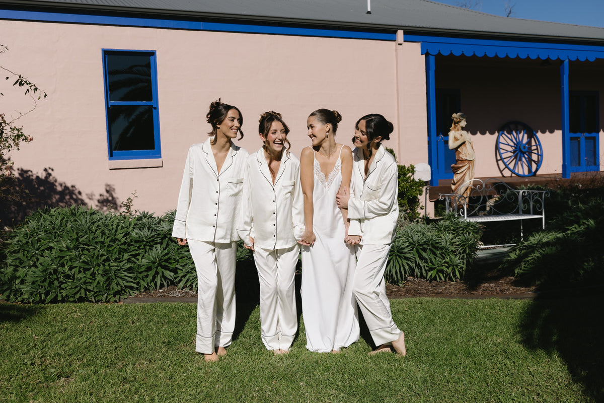 Bride in an ivory satin slip nightgown walking with three bridesmaids in matching ivory satin pyjama sets on the wedding morning