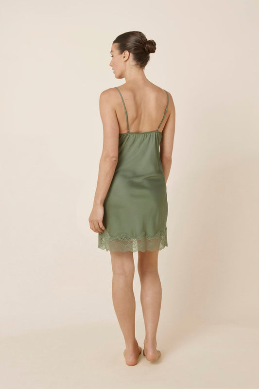 Woman wearing Azalia Silky Satin Short Chemise Olive standing turned away to show the back of the satin slip dress with slim straps and soft flowing fabric.
