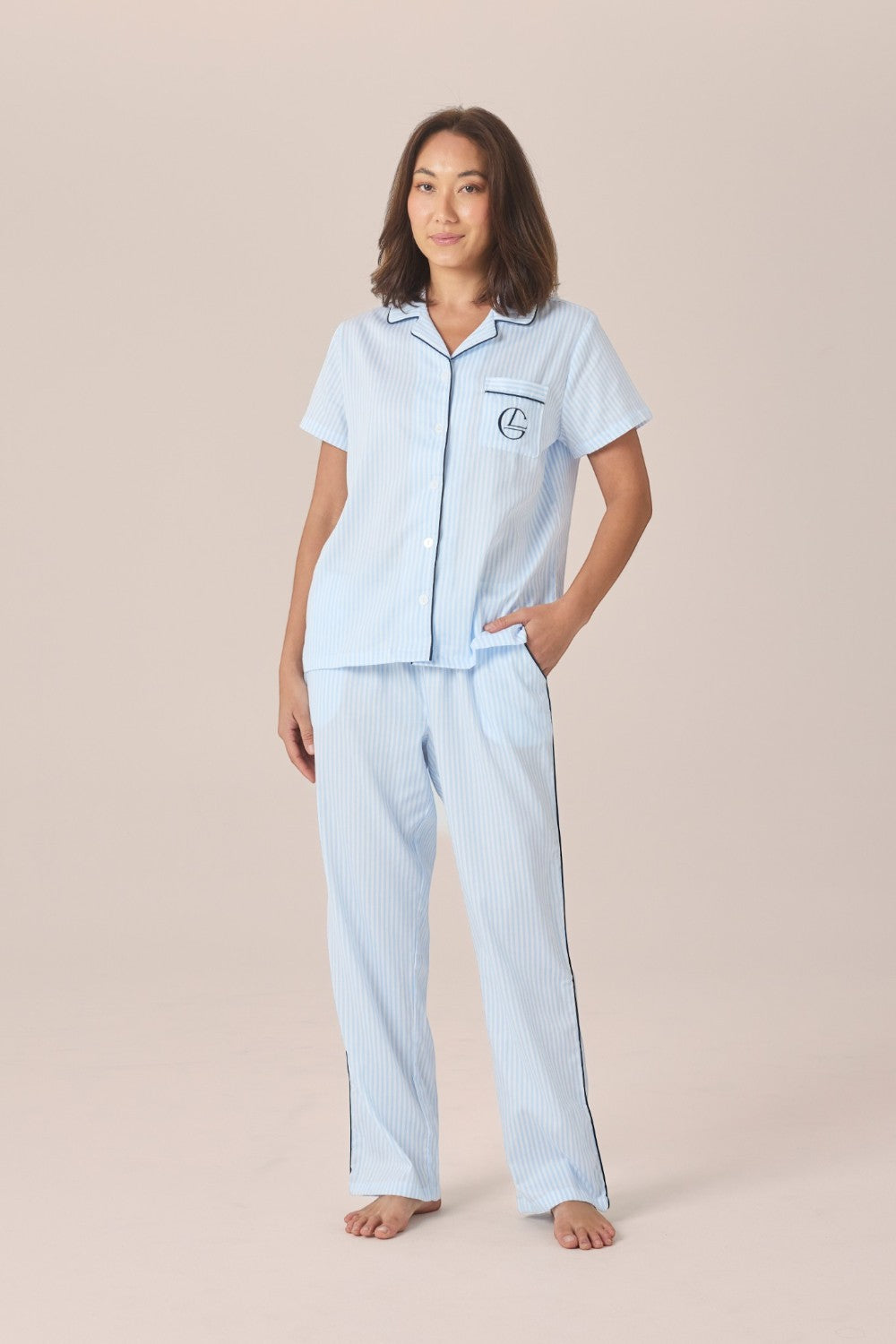 A woman modeling a light blue and white striped pyjama set with a short-sleeve button-up top and long pants, standing with one hand in her pocket against a solid beige studio background.