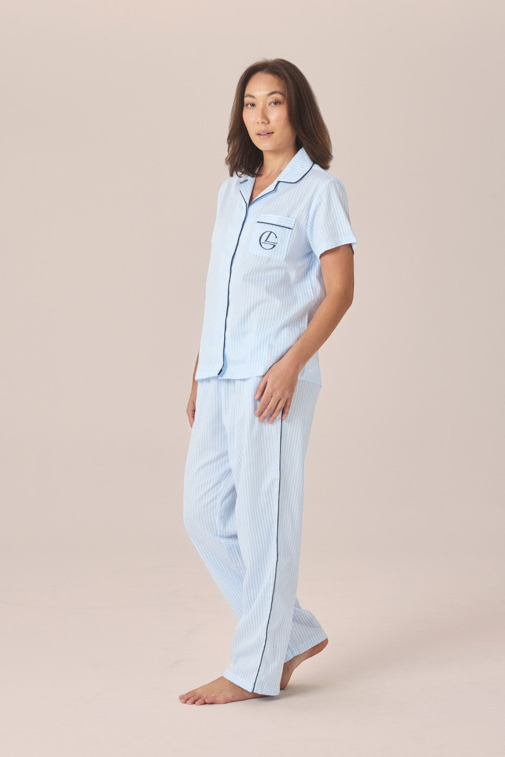 A woman modeling a light blue and white striped pyjama set with a short-sleeve top and long pants, one hand resting in the pocket, against a solid beige studio background.