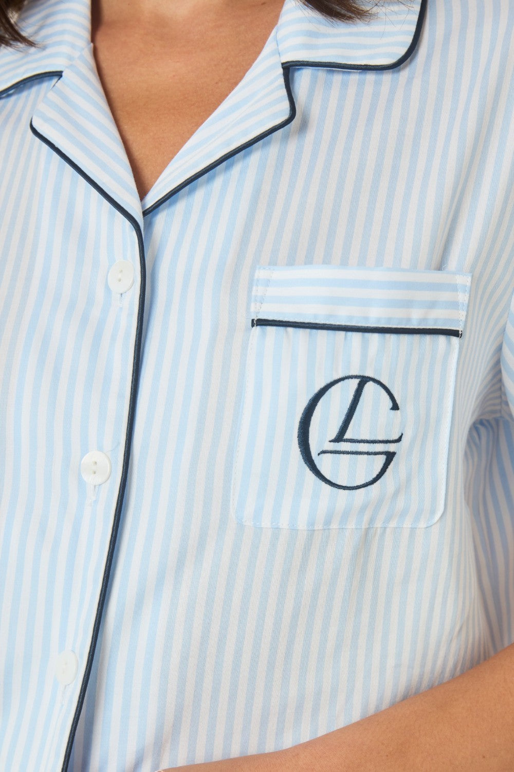Close-up of the collar and embroidered logo pocket on a light blue and white striped pyjama shirt.