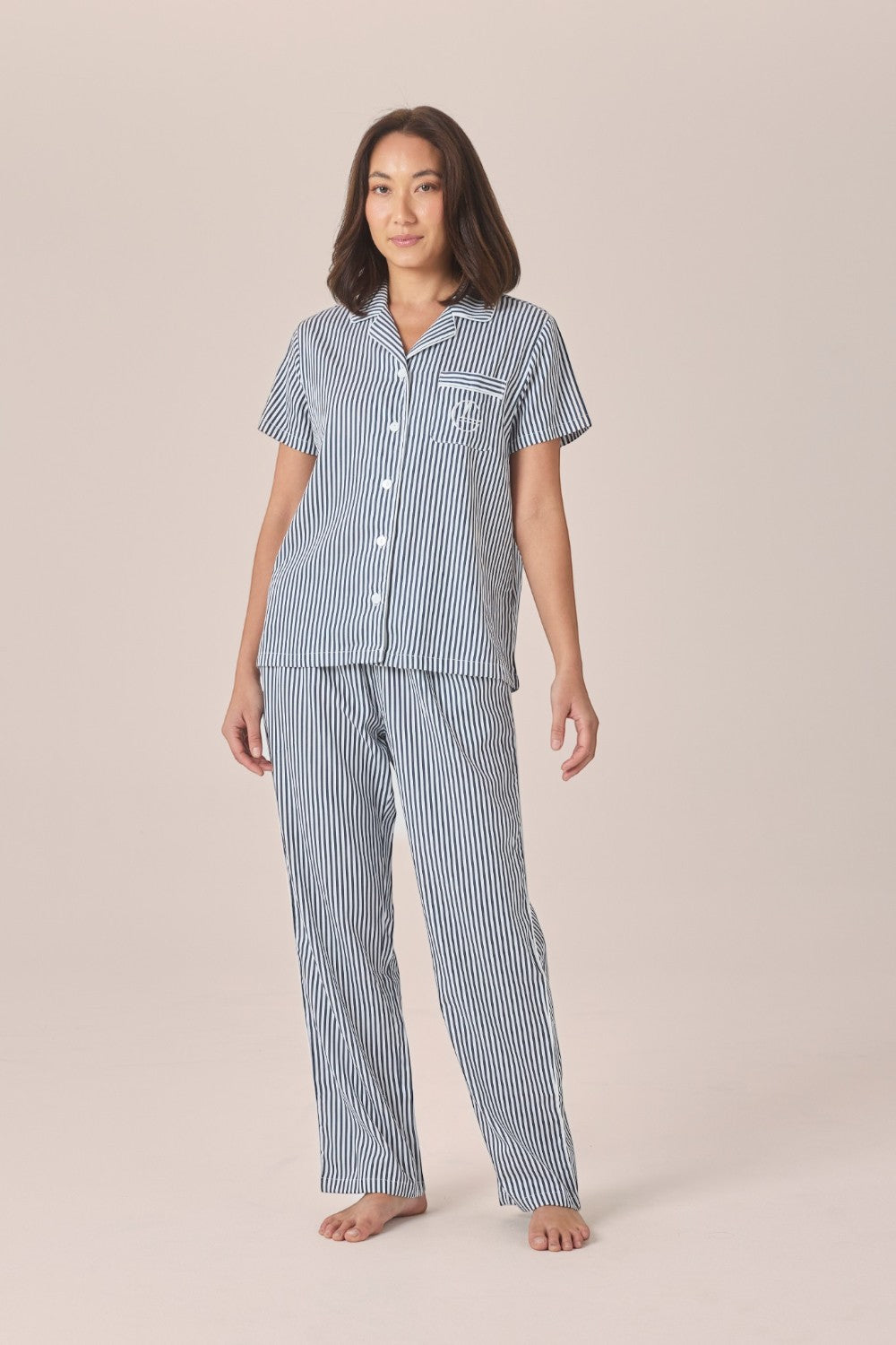 A woman modeling a matching navy and white striped pyjama set with a short-sleeve button-front top and long pants, against a solid beige studio background.