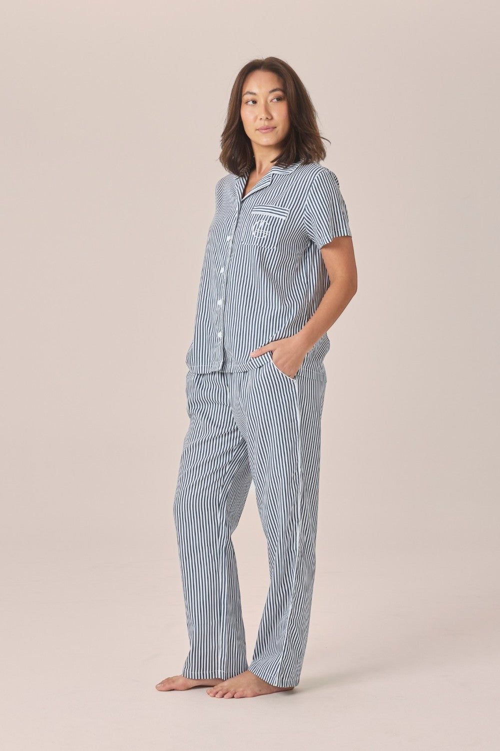 A woman modeling a navy and white striped pyjama set with a short-sleeve top and long pants, one hand resting in the pocket, against a solid beige studio background.