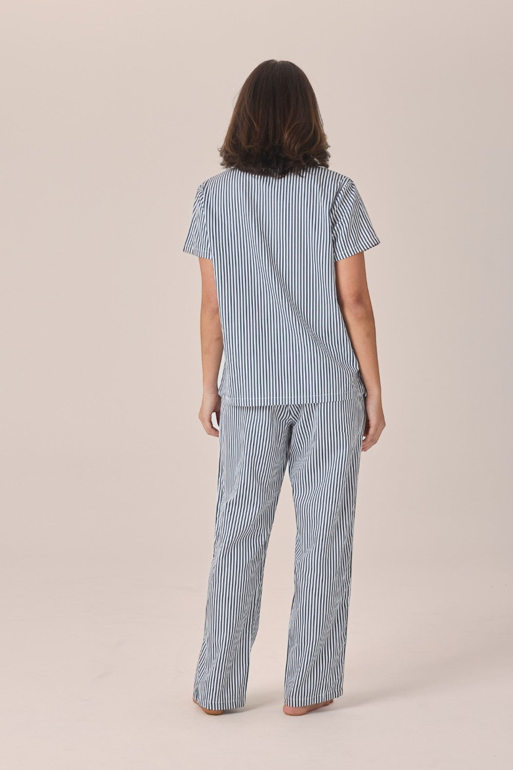 A woman viewed from behind wearing a navy and white striped pyjama set with a short-sleeve top and long pants, standing barefoot against a plain beige studio background.