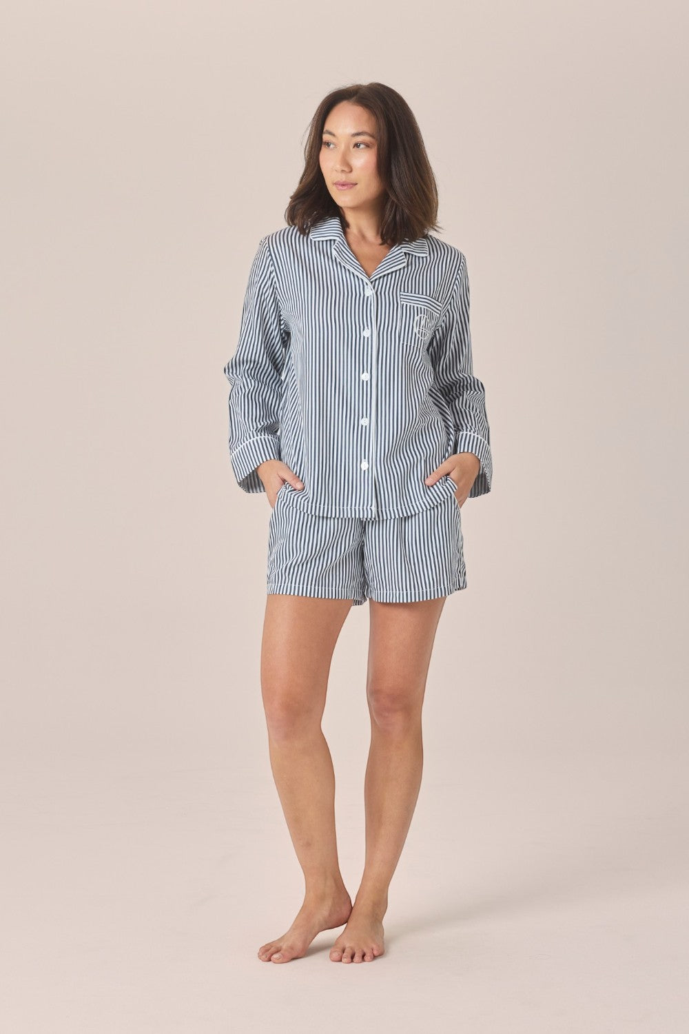 A woman modeling a matching two-piece pajama set with a light blue and white vertical stripe pattern, consisting of a long-sleeve button-up shirt and shorts, against a solid beige studio background.
