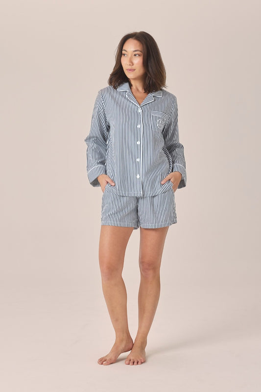 A woman modeling a matching two-piece pajama set with a light blue and white vertical stripe pattern, consisting of a long-sleeve button-up shirt and shorts, against a solid beige studio background.