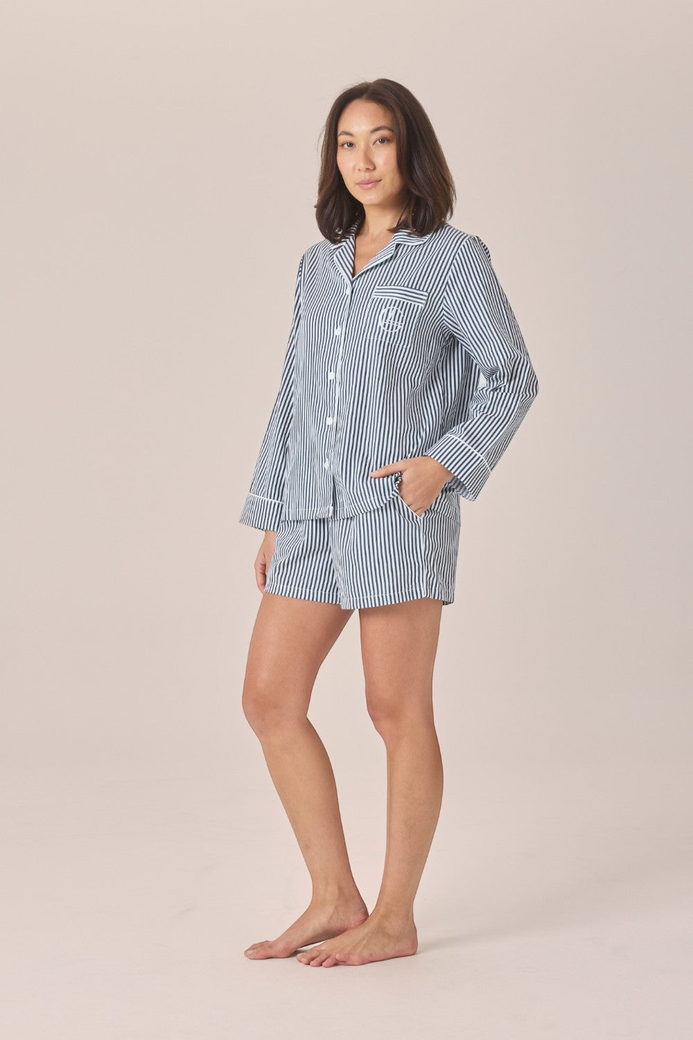 A woman modeling a matching two-piece pajama set with light blue and white vertical stripes, consisting of a long-sleeve button-up shirt and matching shorts, against a solid beige studio background.