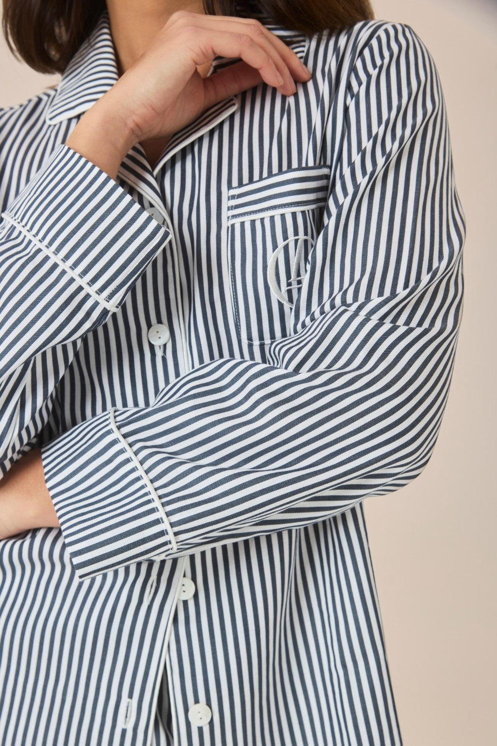 Close-up of the sleeve and embroidered logo pocket on a navy and white striped pajama shirt, worn by a woman with her hand raised to her collar.