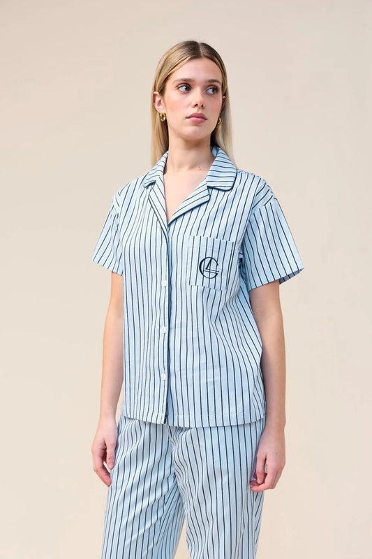 A woman modeling a matching short-sleeve button-front pajama top and striped pajama pants set in light blue and white. The top features a chest pocket with a circular embroidered logo.