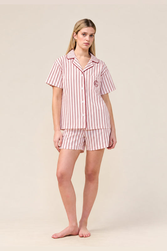 A woman modeling a matching two-piece pajama set with short-sleeve button-front top and shorts, featuring vertical pink and white stripes with a small embroidered crest on the chest, against a plain beige studio background.