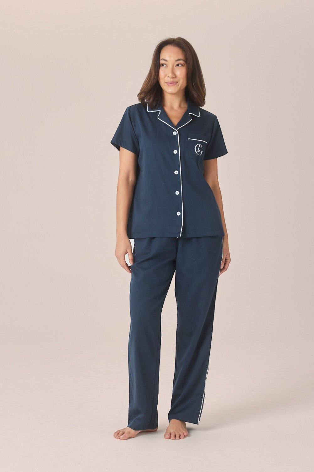 A woman modeling a matching navy pyjama set with a short-sleeve button-front top and long pants, featuring a white contrast piping and embroidered logo pocket, against a solid beige studio background.
