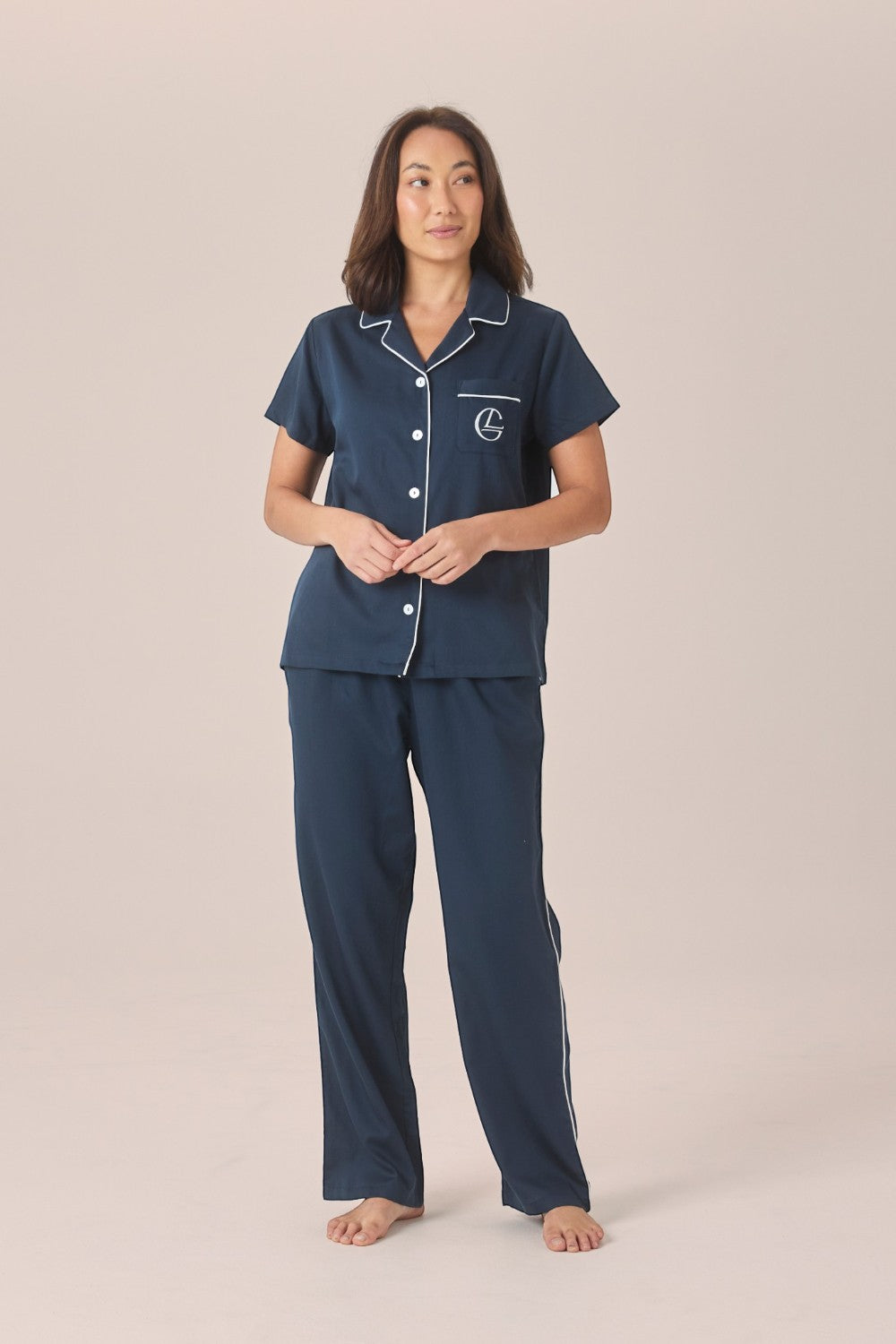 A woman modeling a navy pyjama set with a short-sleeve button-up top and long pants, standing with hands clasped against a solid beige studio background.