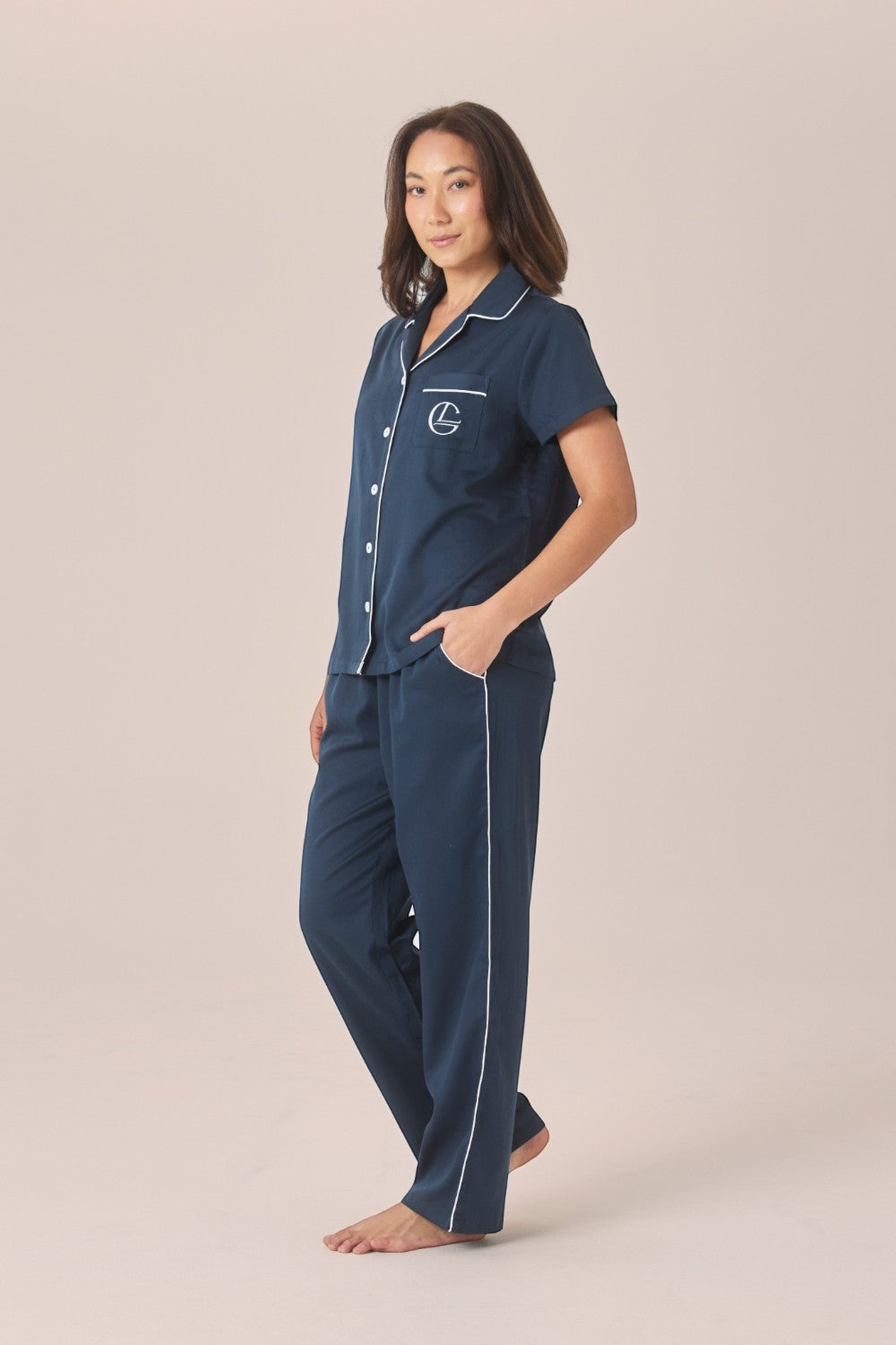 A woman modeling a navy pyjama set with a short-sleeve top and long pants, one hand resting in the pocket, against a solid beige studio background.