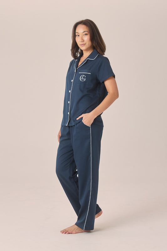 A woman modeling a navy pyjama set with a short-sleeve top and long pants, one hand resting in the pocket, against a solid beige studio background.