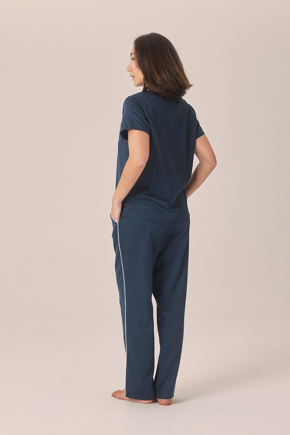 A woman viewed from behind wearing a navy pyjama set with a short-sleeve top and long pants, looking back over her shoulder with one hand in her pocket.