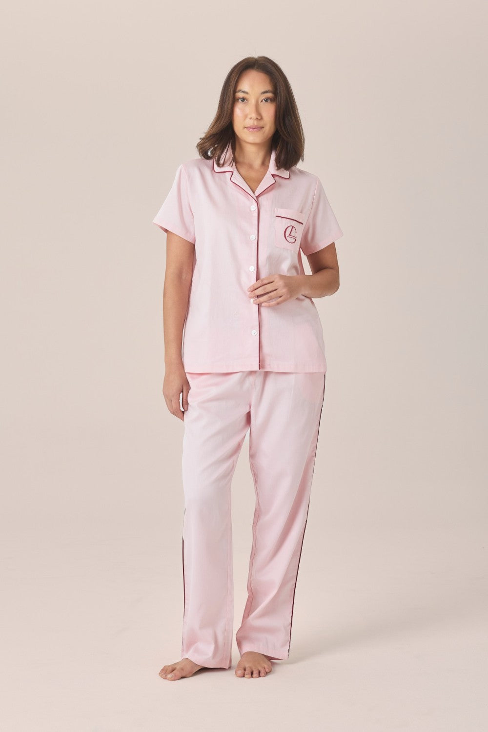 A woman modeling a pale pink pyjama set with a short-sleeve button-up top and long pants, standing against a solid beige studio background.