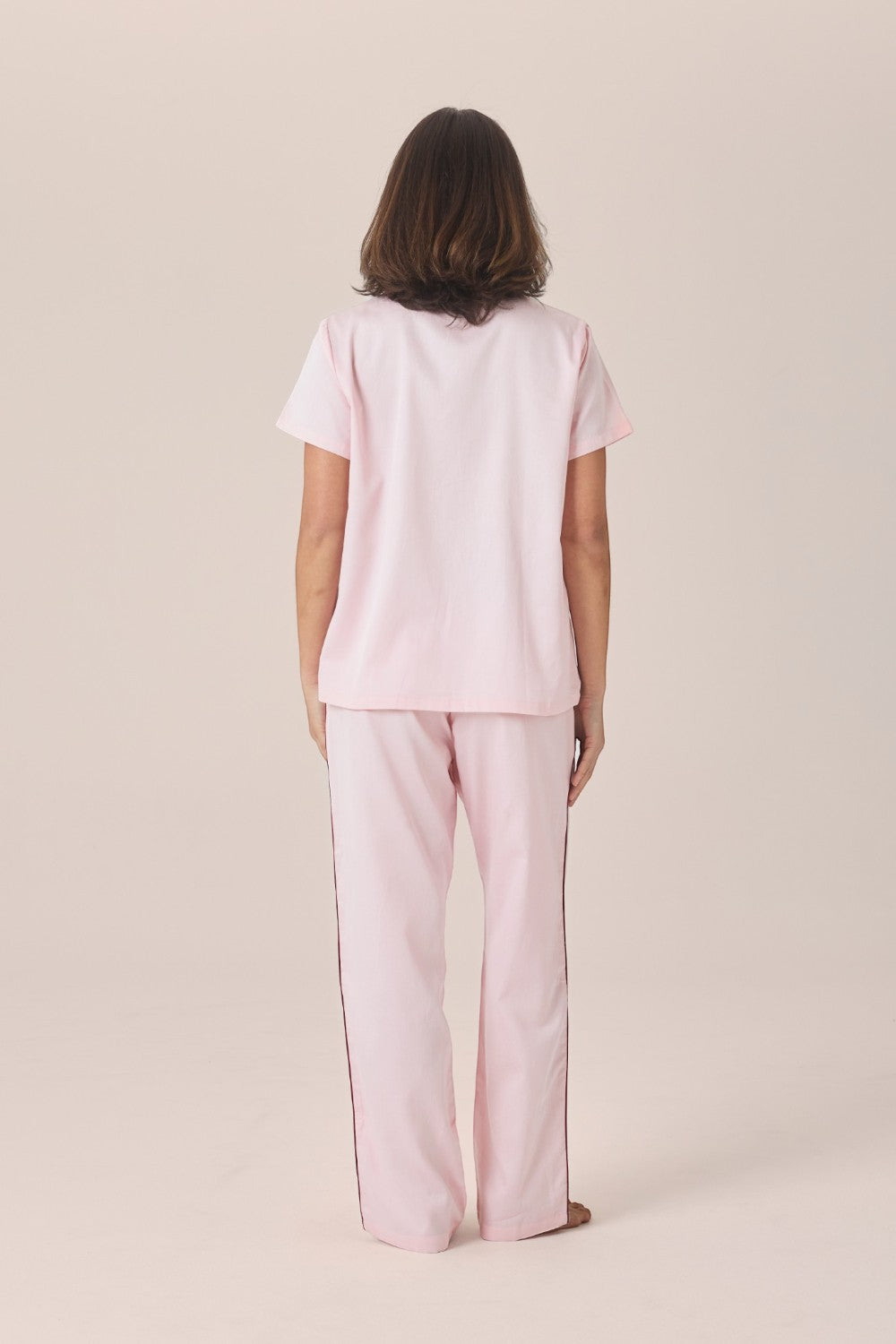 A woman viewed from behind wearing a pale pink pyjama set with a short-sleeve top and long pants, standing barefoot against a plain beige studio background.