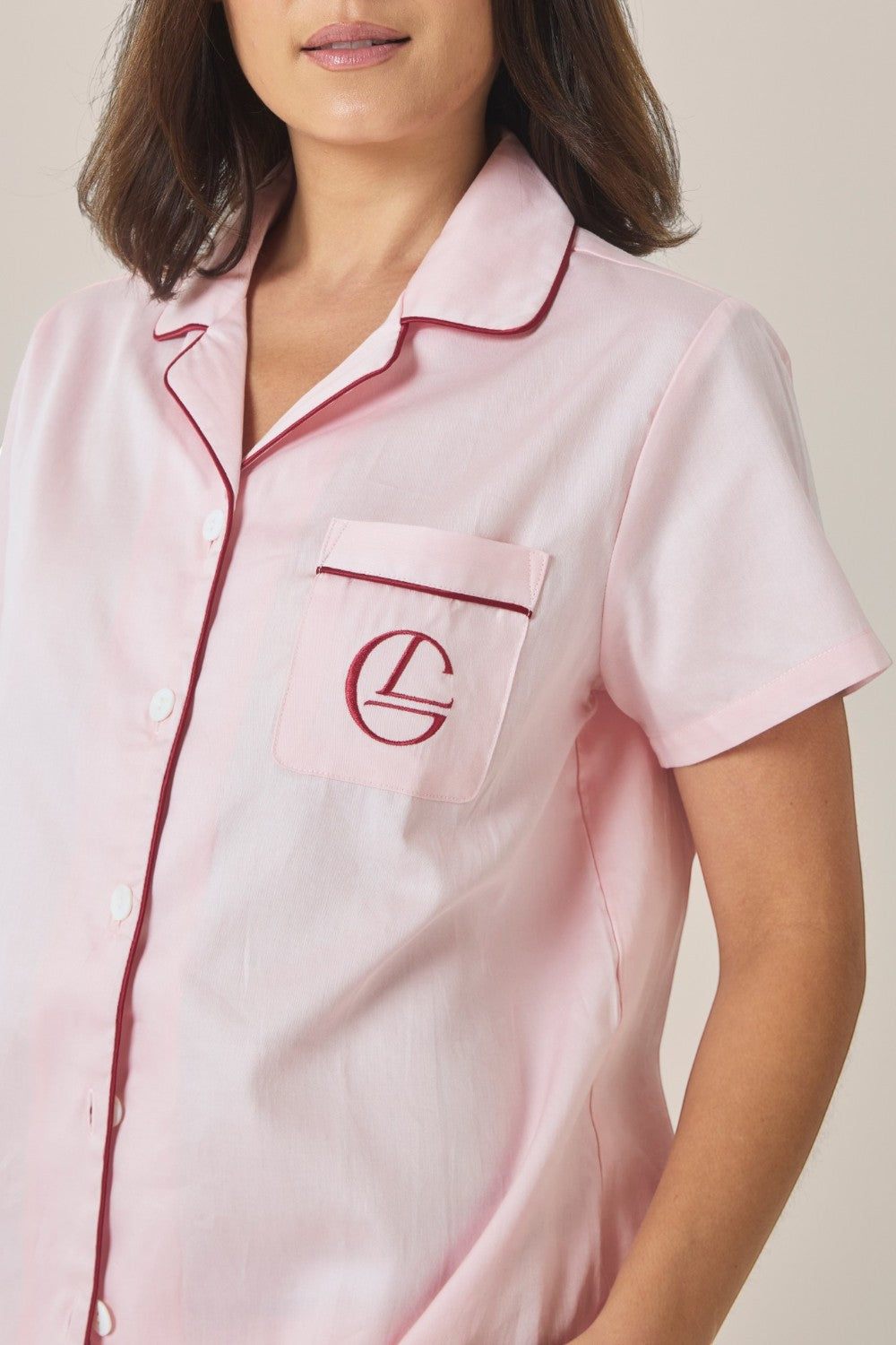 Close-up of the collar and embroidered logo pocket on a pale pink pyjama shirt with contrast burgundy piping.
