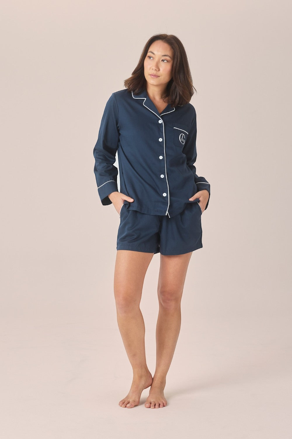 A woman modeling a navy blue pajama set with white piping details, consisting of a long-sleeve button-up shirt and matching shorts, against a solid light beige background.