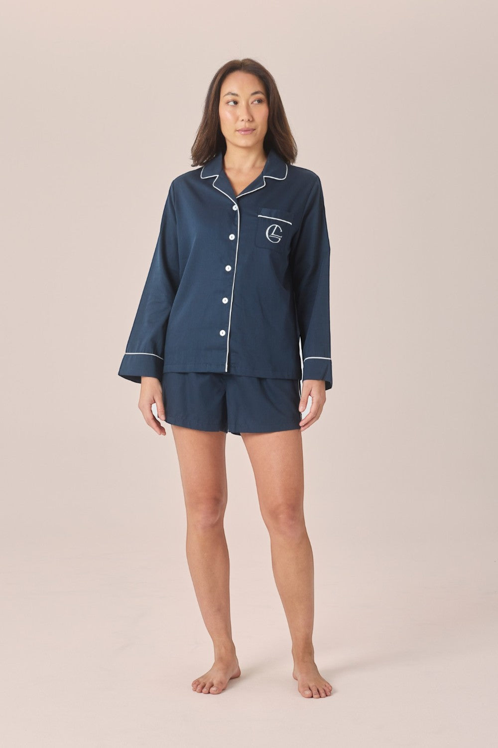 A woman modeling a navy blue pajama set with white piping, consisting of a long-sleeve button-front top and matching shorts, against a solid light beige background.