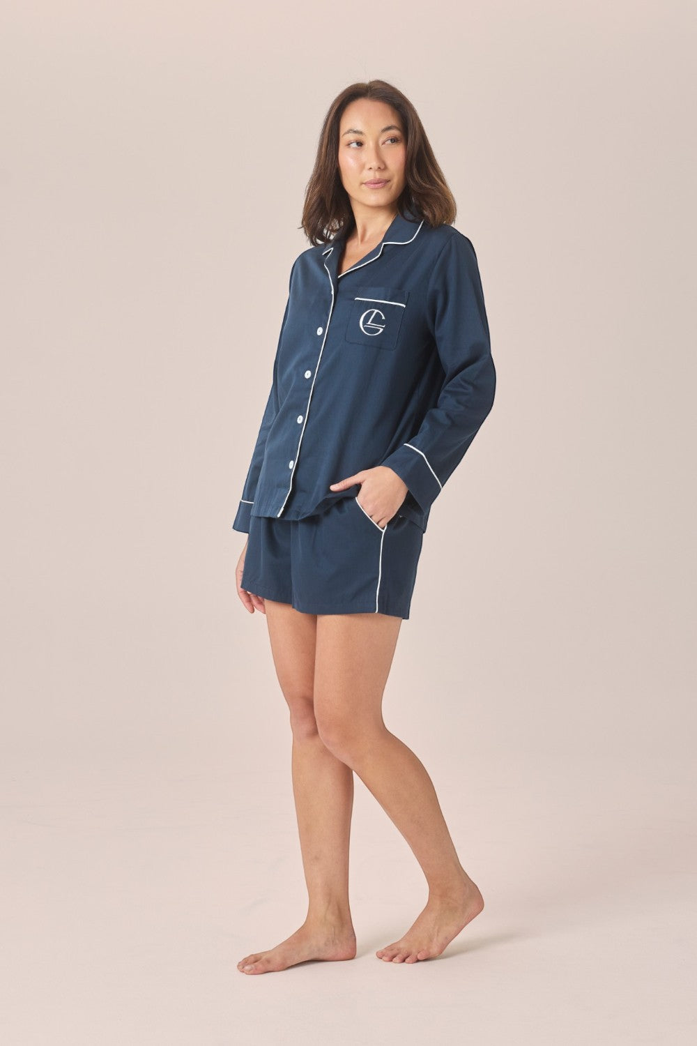 A woman modeling a navy blue pajama set with white piping, consisting of a long-sleeve button-front top and matching shorts, against a solid light beige studio background.