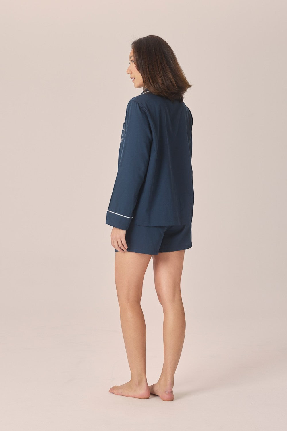A woman stands barefoot in profile wearing a navy blue pajama set with white piping trim, consisting of a long-sleeve top and matching shorts, against a solid light beige background.