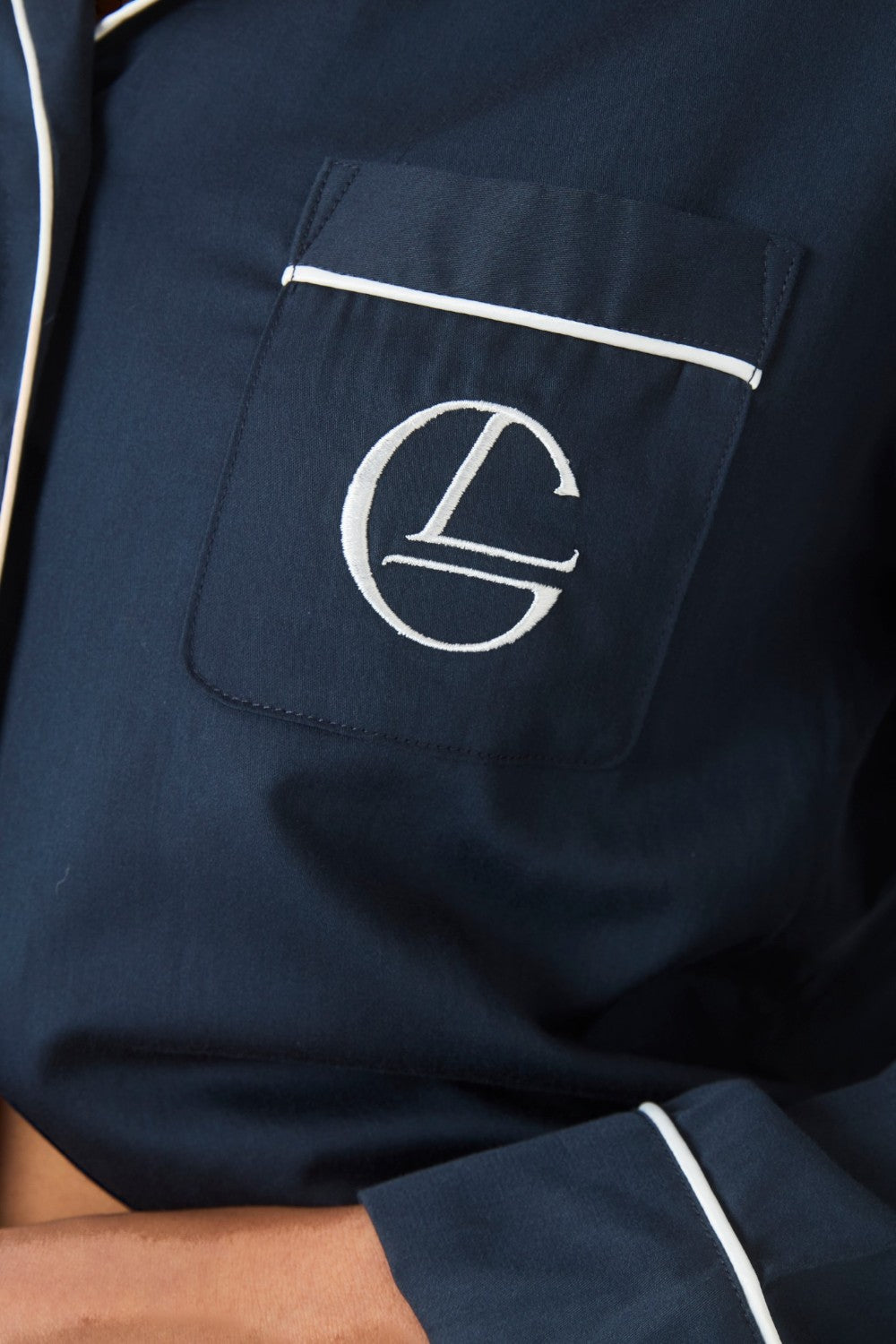 Close up of the embroidered logo on the chest pocket of the Susanna navy pyjama top, with white piping detail along the pocket edge.
