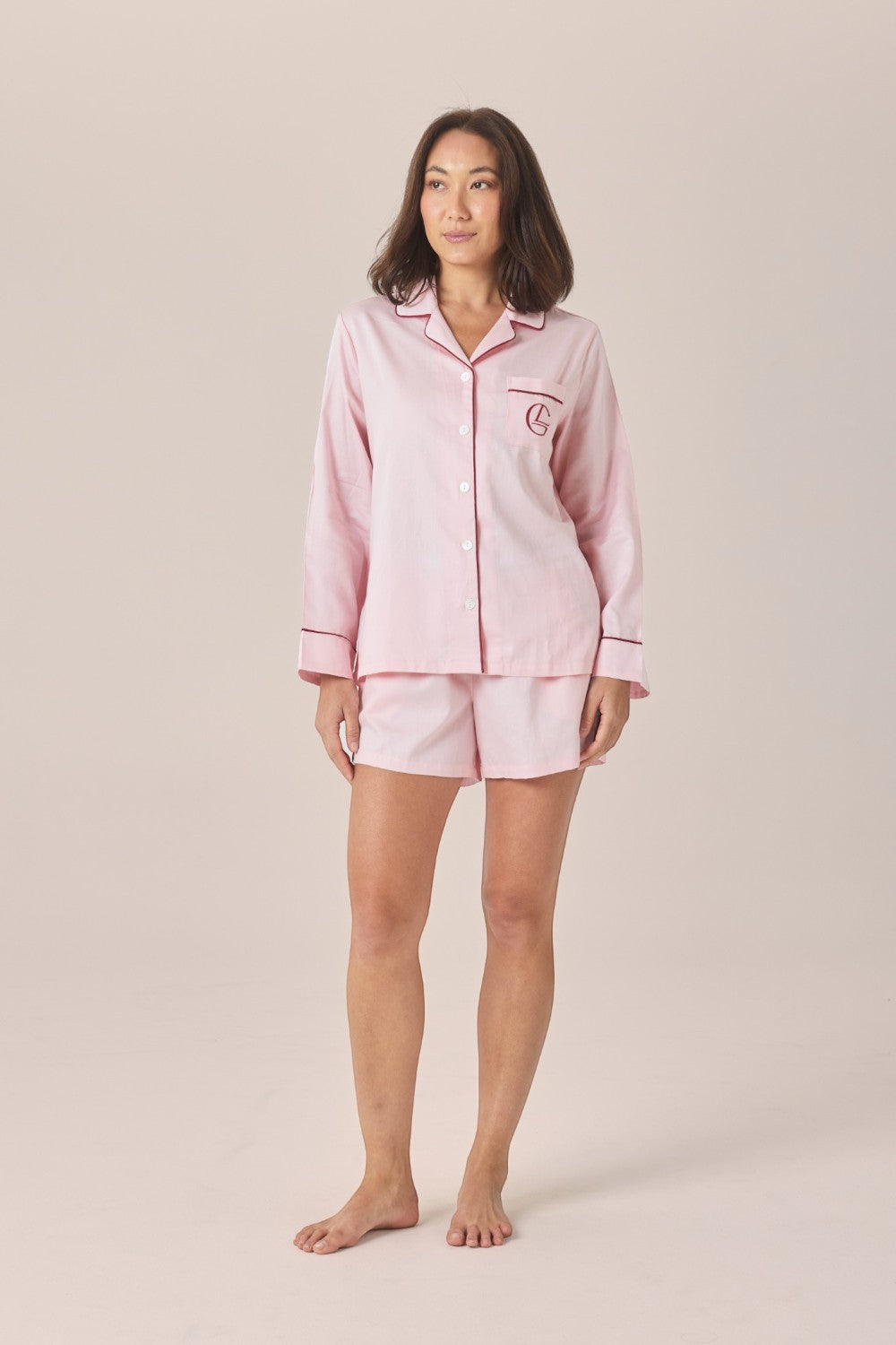 A woman modeling a matching two-piece pajama set in pale pink with a long-sleeve button-front top and shorts, featuring an embroidered logo pocket, against a solid beige studio background.