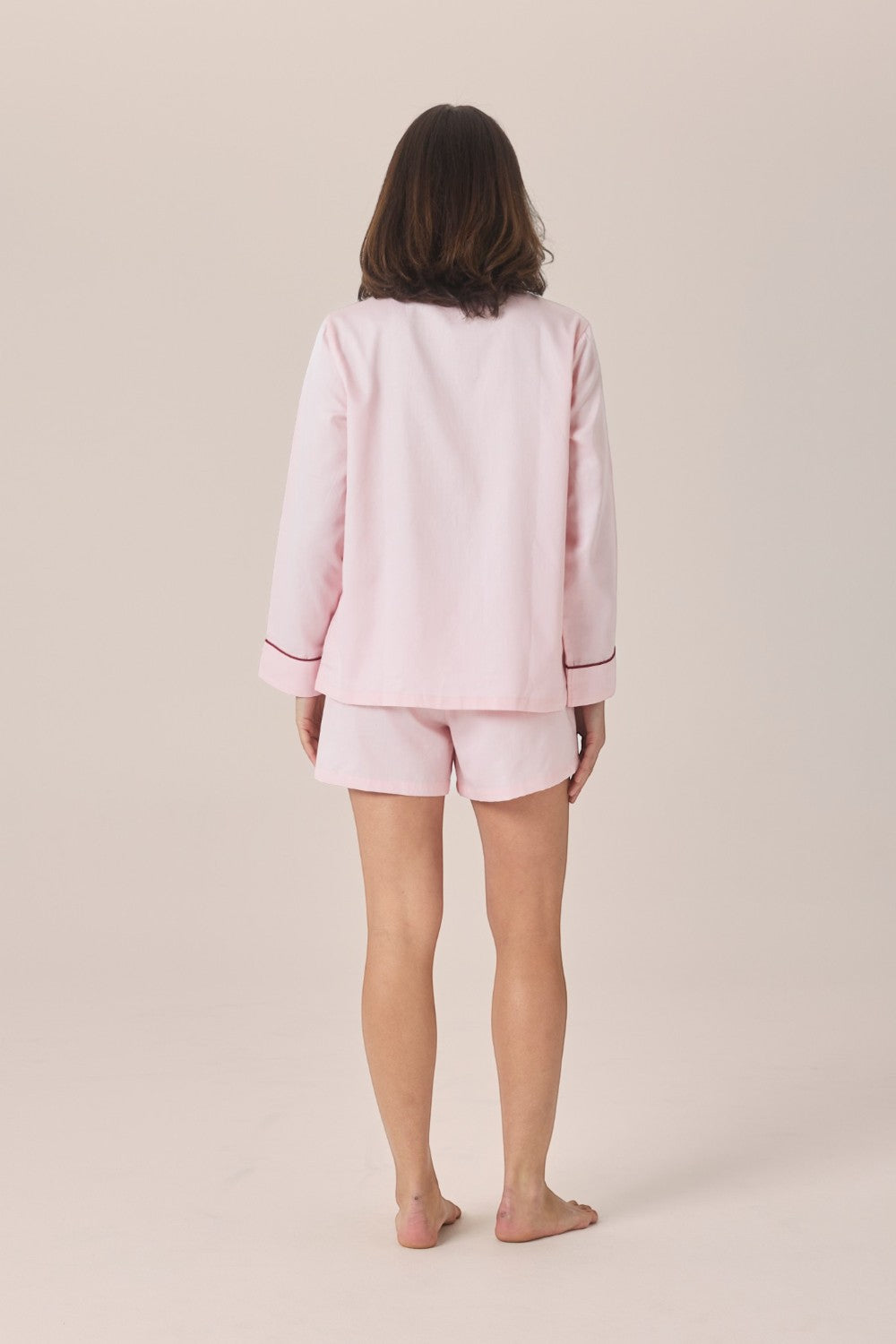 A woman viewed from behind wearing a pale pink pajama set with a long-sleeve top and matching shorts, standing barefoot against a plain beige studio background.