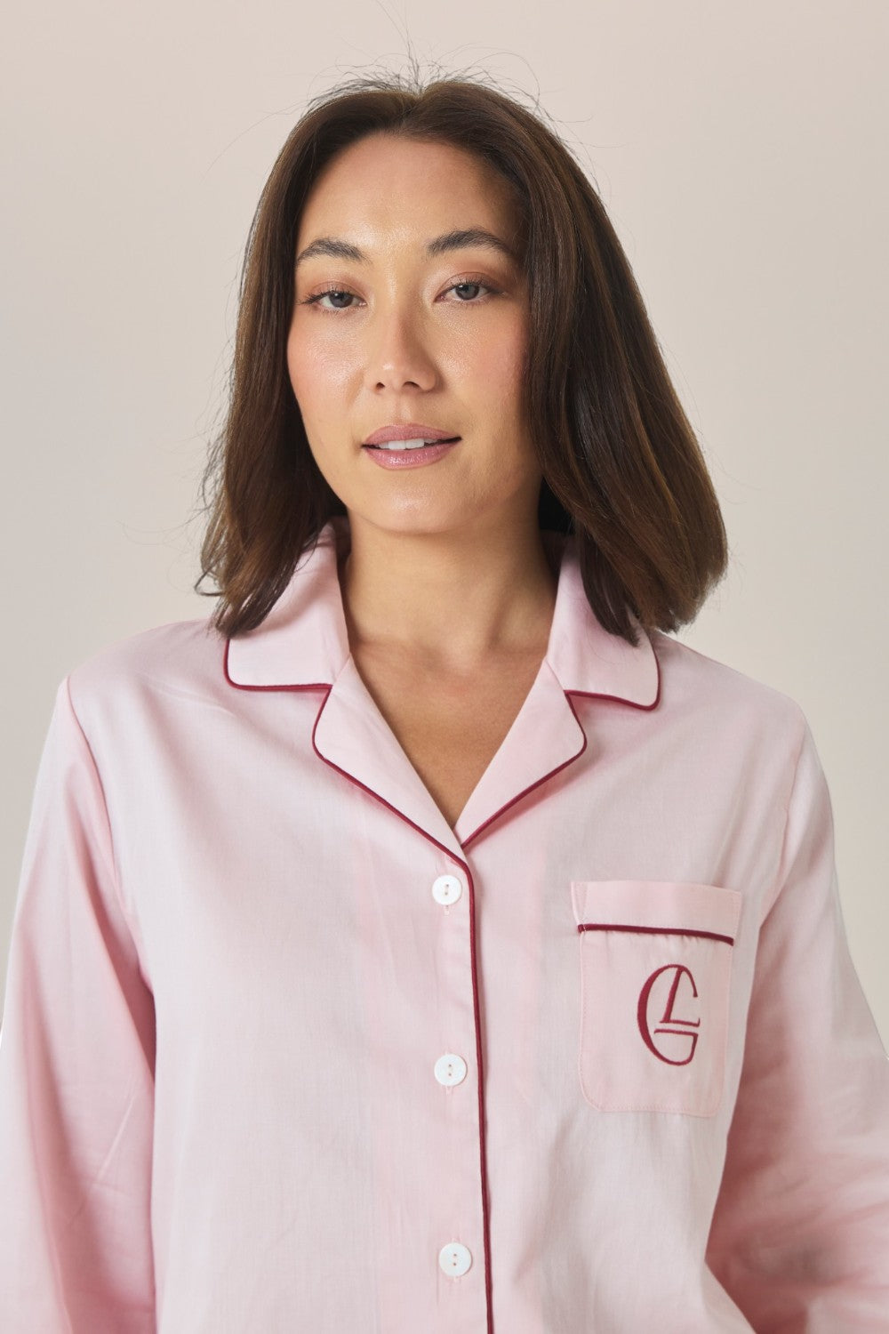 Close-up of a woman wearing a pale pink pajama shirt with contrast burgundy piping and an embroidered logo pocket.
