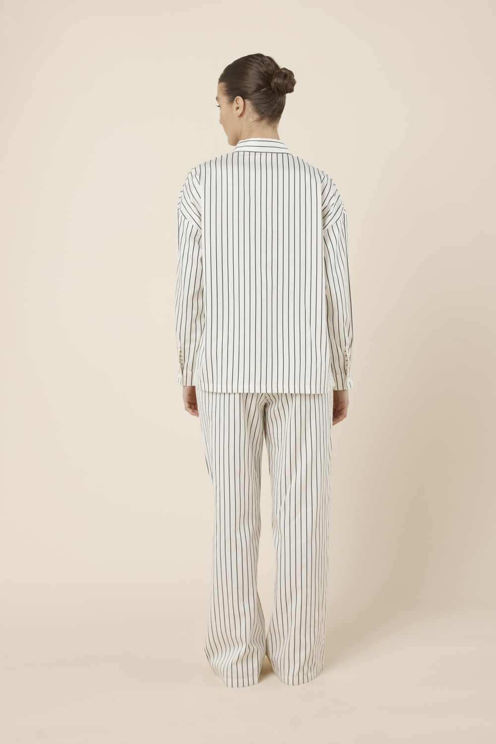 Woman wearing a black and white striped ivory willow cotton stripe pyjama PJ set in set on a beige background back view