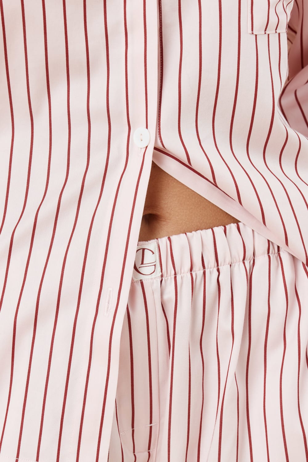 Close up waistline view of woman wearing a pink striped willow cotton stripe pyjama PJ set on a beige background showing the gingerlilly logo and white button