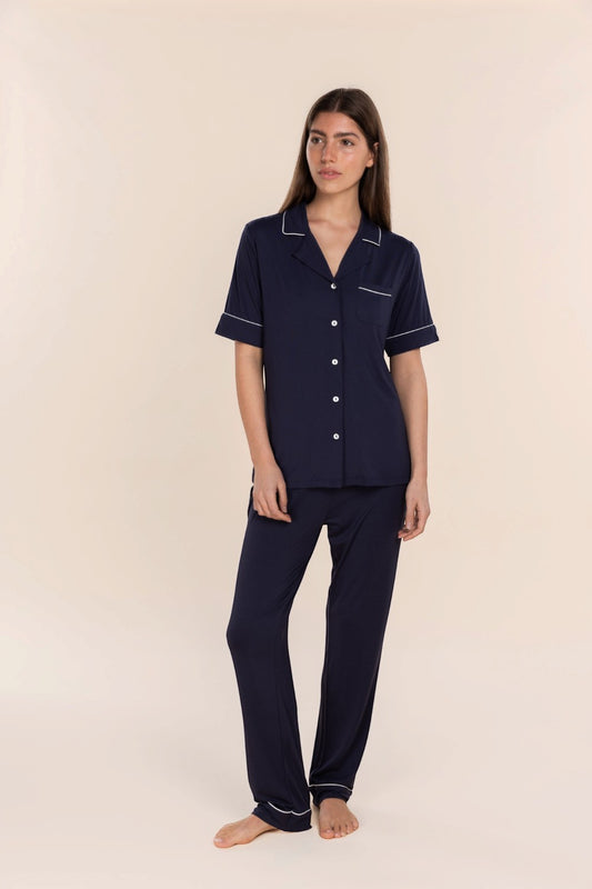 A woman modeling a navy blue two-piece pajama set with white piping details, consisting of a short-sleeve button-up shirt and matching pants, against a solid light beige background.