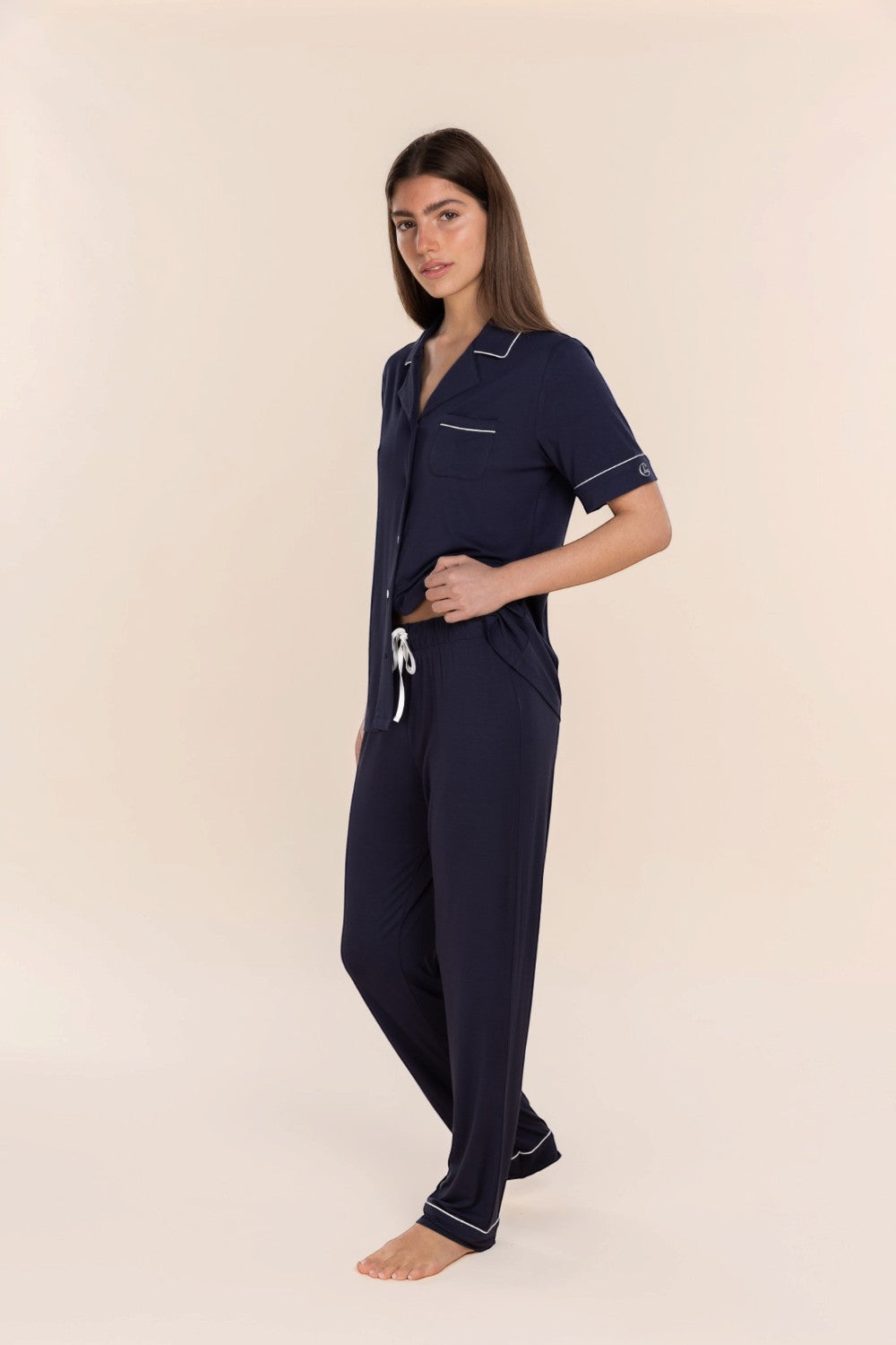 A woman modeling a navy blue short-sleeve pajama set with white piping and a white drawstring, standing against a solid light beige background.