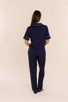 A woman viewed from the back wearing a navy blue short-sleeve pajama top and matching pants with thin white piping trim, standing against a plain beige studio background.