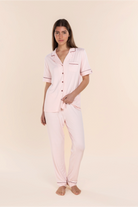 A woman modeling a light pink pajama set consisting of a short-sleeve button-up top and matching pants, with thin dark pink piping details on the collar, pocket, and cuffs, set against a solid pale beige background.