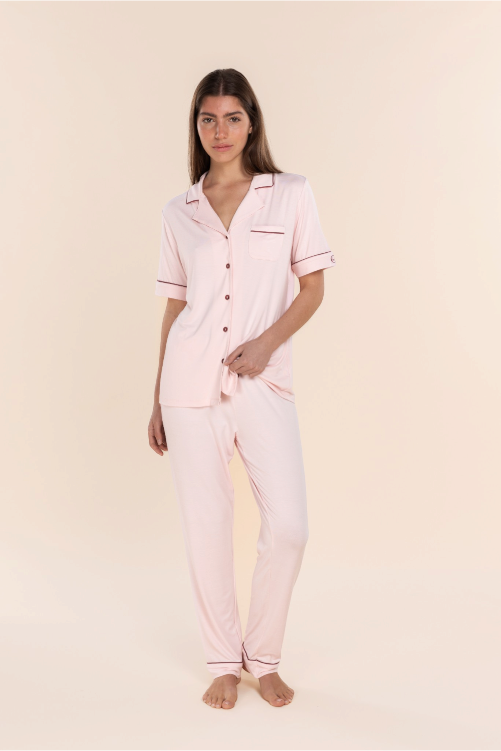 A woman modeling a light pink pajama set consisting of a short-sleeve button-up top and matching pants, with thin dark pink piping details on the collar, pocket, and cuffs, set against a solid pale beige background.