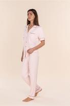 A woman modeling a light pink pajama set with thin dark pink piping details, consisting of a short-sleeve button-up shirt and matching pants, against a solid cream background.